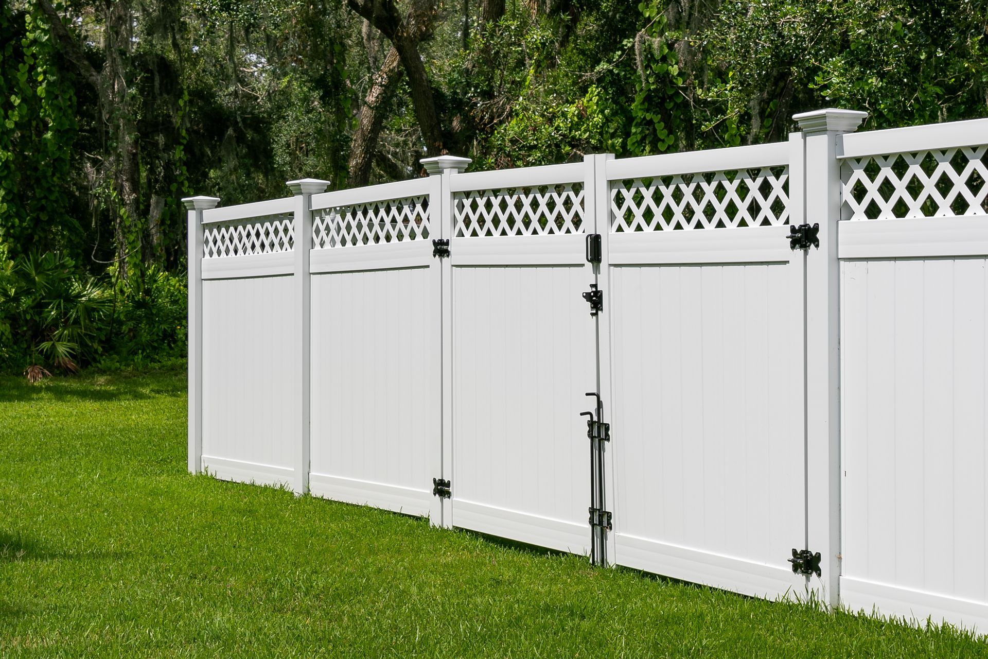 A white vinyl fence with a lattice top and a double gate, set against a green lawn and trees.