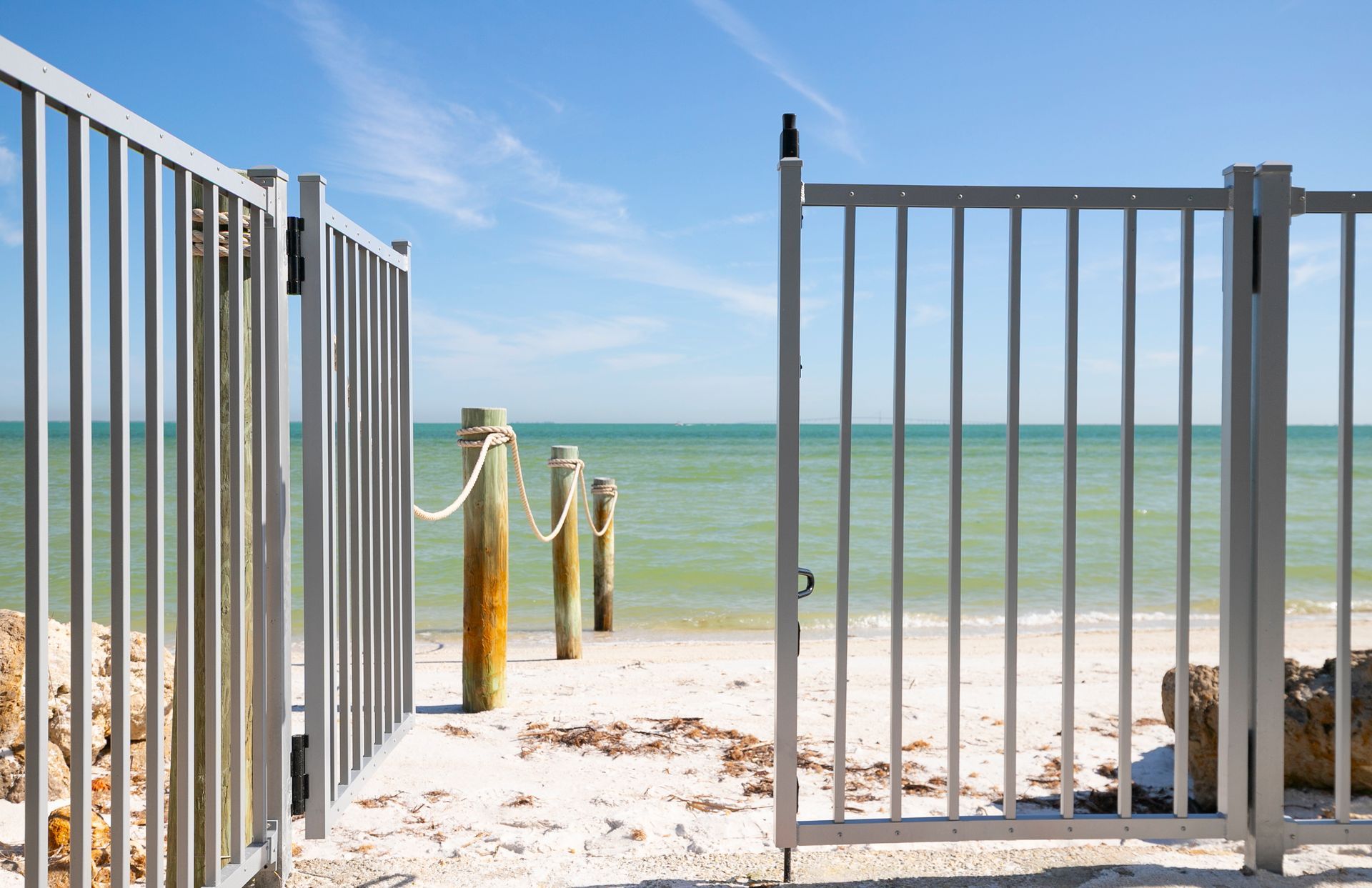 An open metal gate leads to a sandy beach with wooden posts and a rope fence overlooking the ocean under a blue sky.