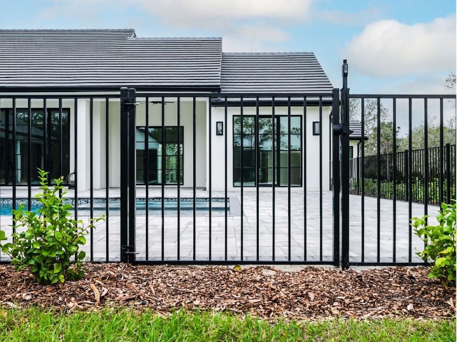 A black metal pool fence stands in front of a modern white house with a patio and swimming pool.