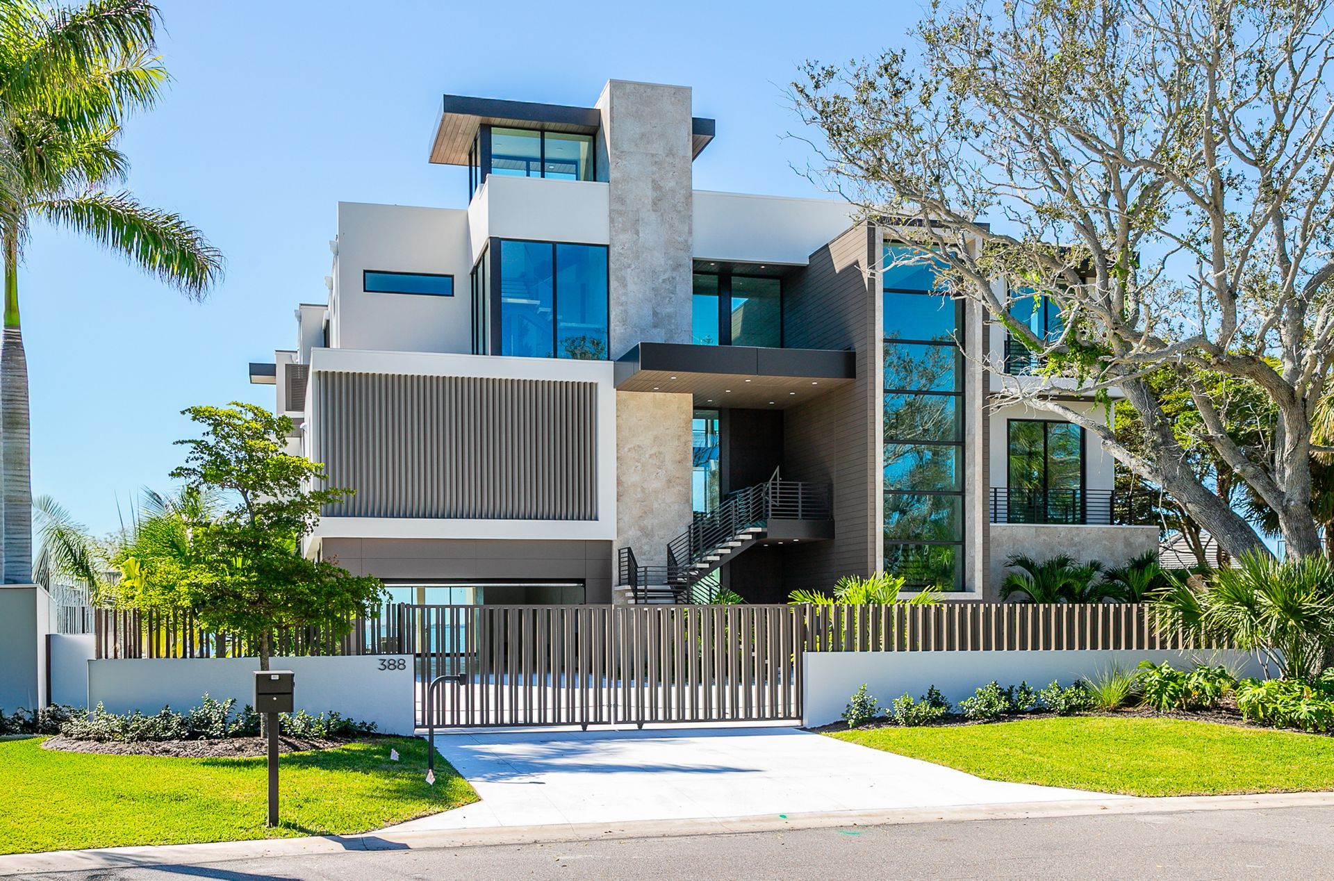 A modern, three-story house with light stone accents, large windows, and a slatted front gate, surrounded by green trees.