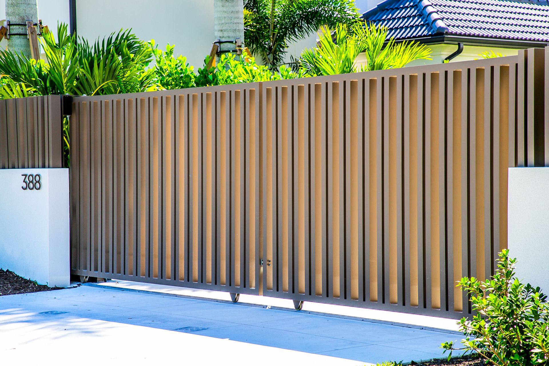 A wide, tan, vertical-slat metal sliding gate stands between two white pillars, with lush green plants in the background.