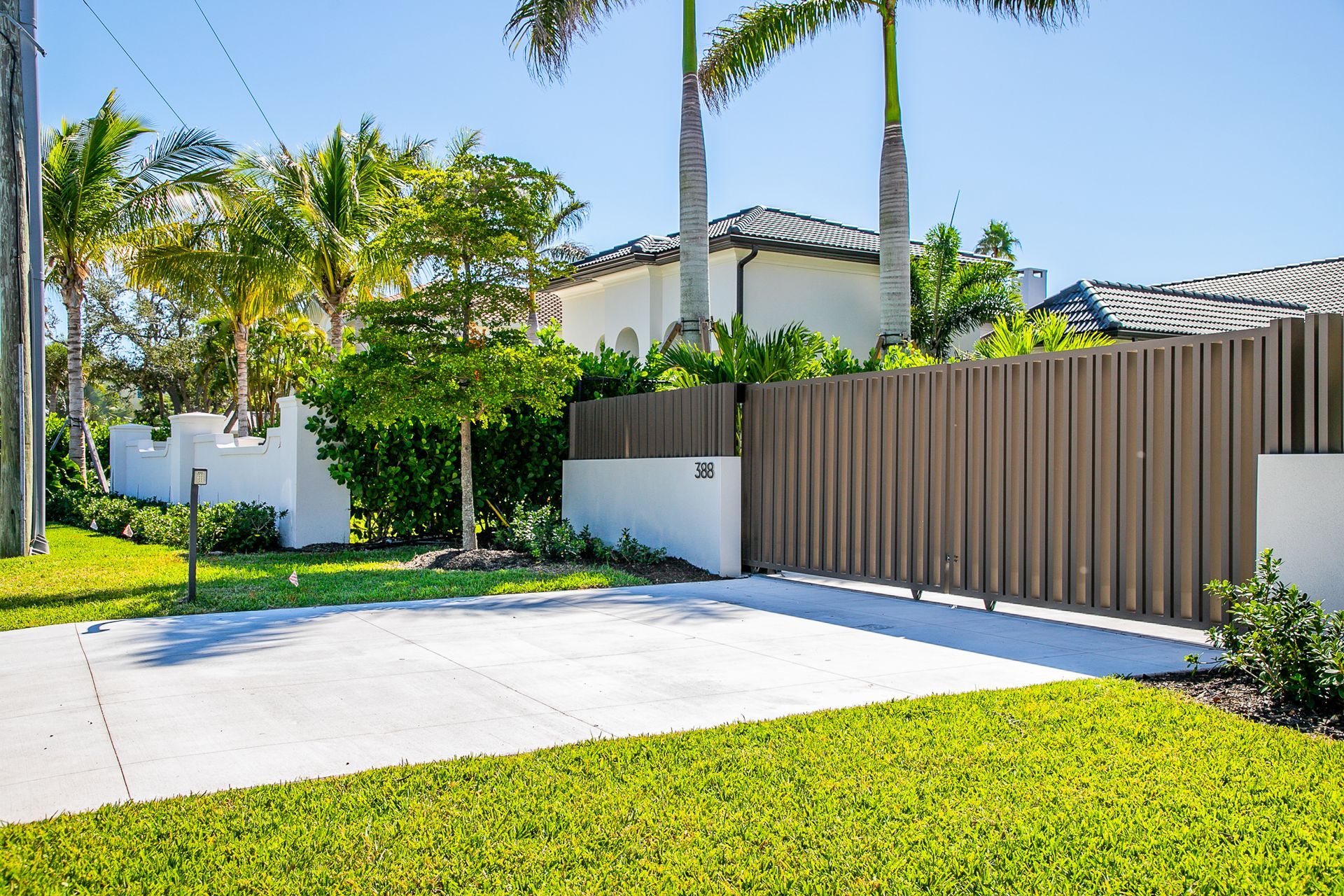 A concrete driveway leads to a gated entrance with a tall, wooden fence and white masonry walls, surrounded by palm trees.