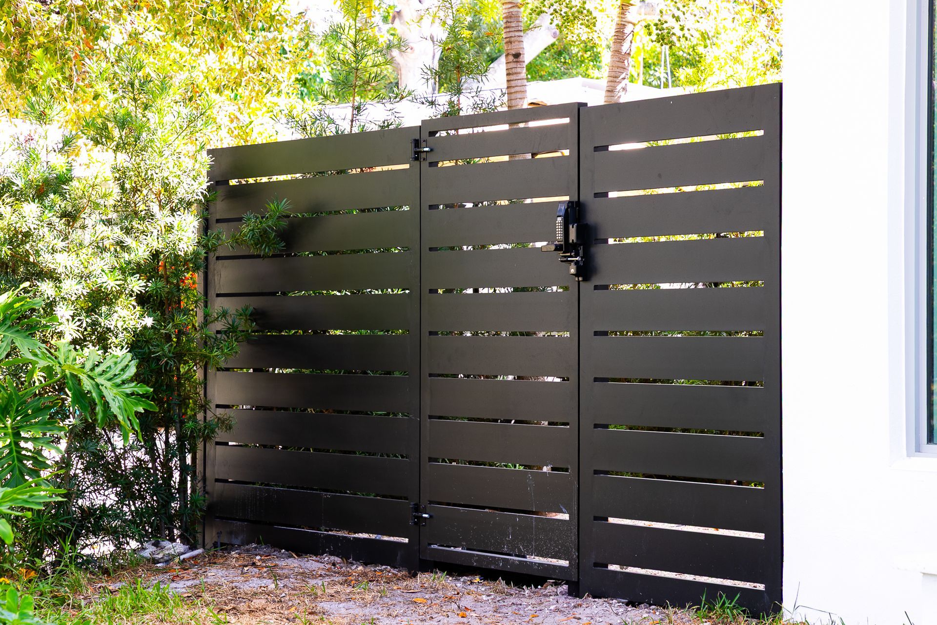 A dark brown horizontal slat privacy gate standing outdoors against a white wall and lush greenery.