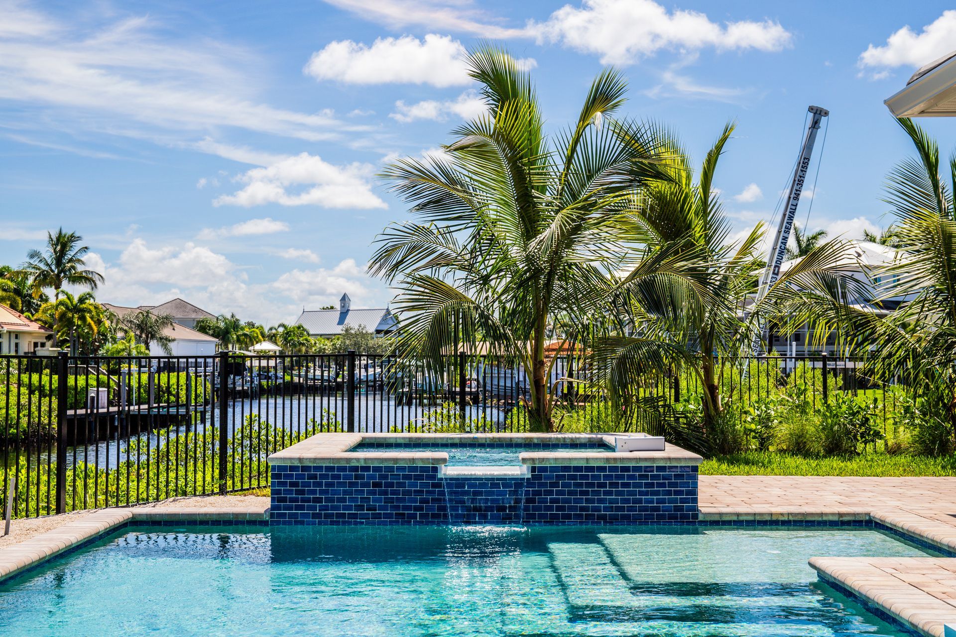 A swimming pool with an attached blue-tiled spa overlooking a waterfront canal surrounded by tropical palm trees.