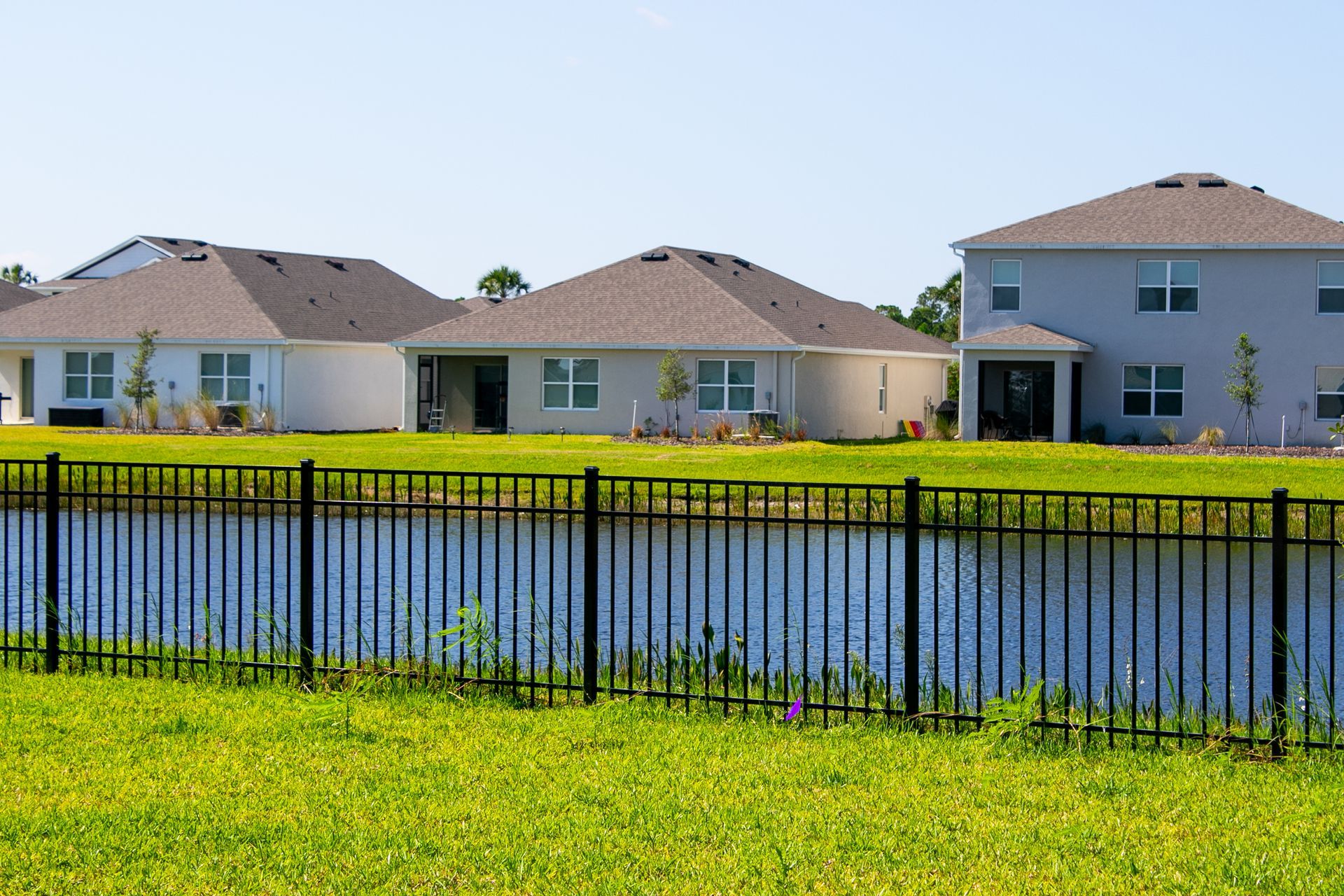 A metal fence stands before a pond, with several suburban houses in the background under a clear blue sky.