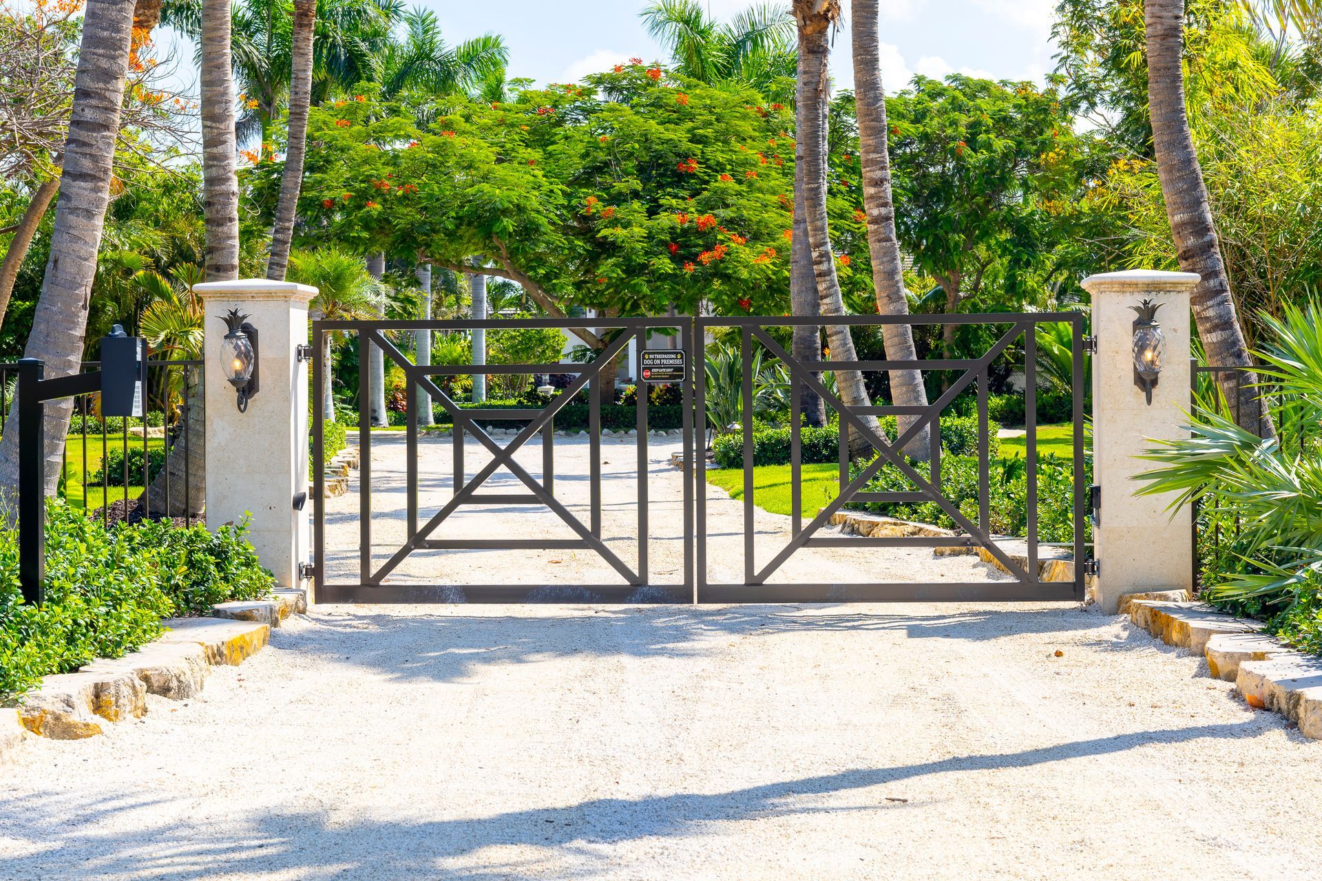 A gated entrance with a metal fence and stone pillars leading to a gravel driveway surrounded by lush palm trees.