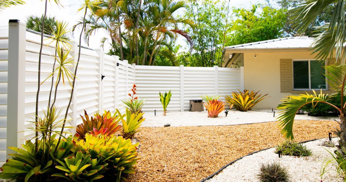 A sunny backyard featuring white horizontal fencing, a tan house, tropical plants, and a gravel pathway.