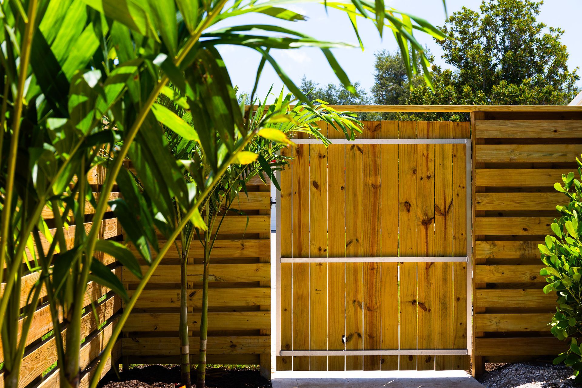 A wooden gate set into a horizontal slat fence, framed by lush green plants in a bright, outdoor setting.