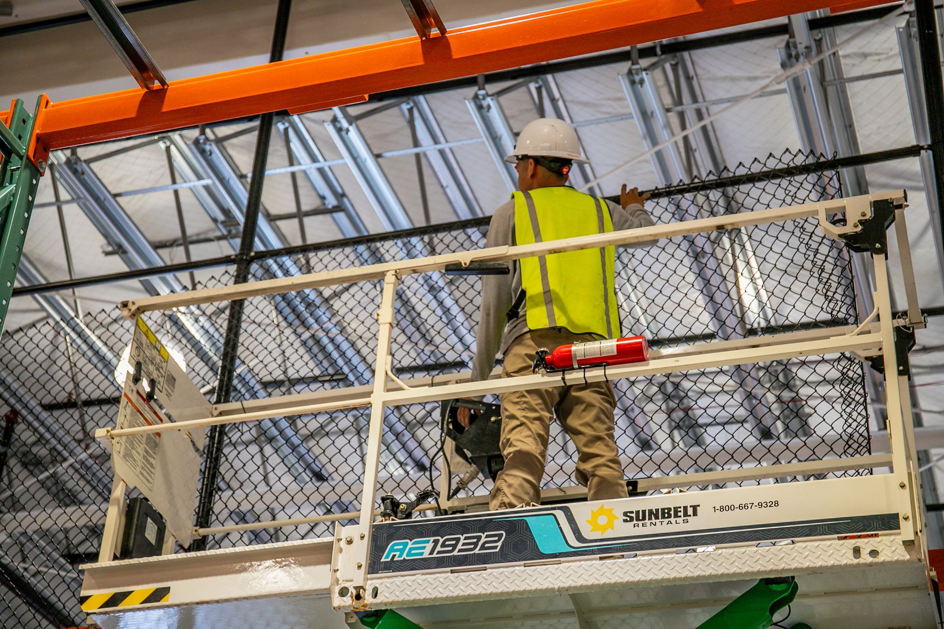 A worker in a high-visibility vest and hard hat stands on an elevated scissor lift, reaching toward industrial ceiling.