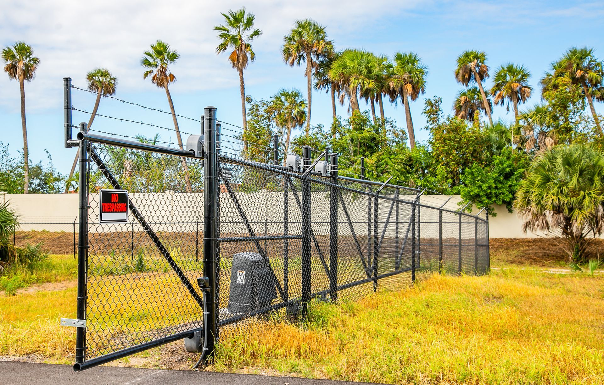 A black chain-link fence with a closed gate, barbed wire top, and warning sign, set against palm trees and dry grass.