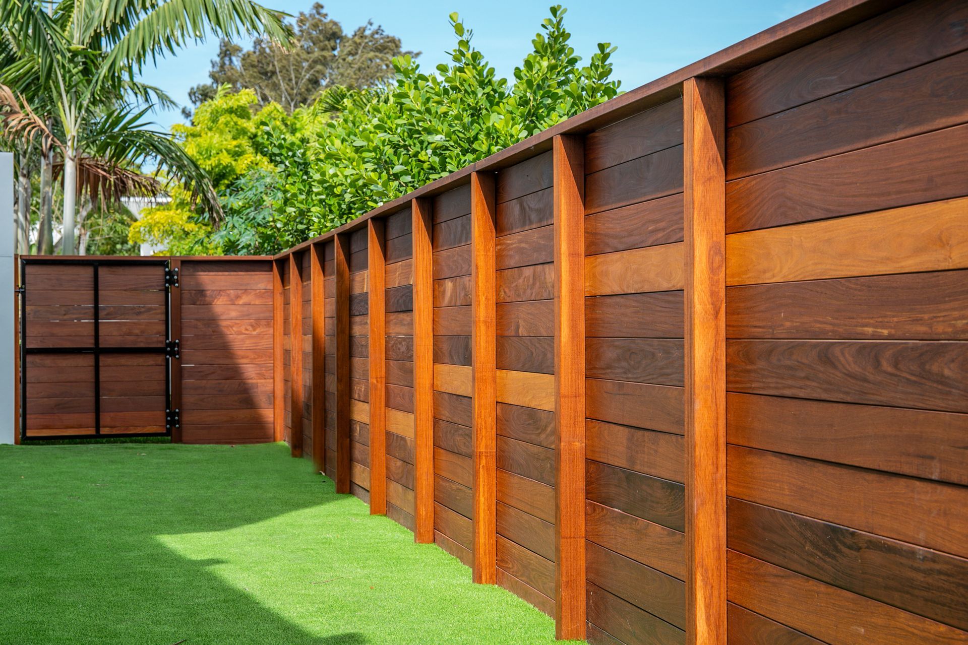 A modern, horizontal wooden slat fence borders a green grassy yard under a bright blue sky.