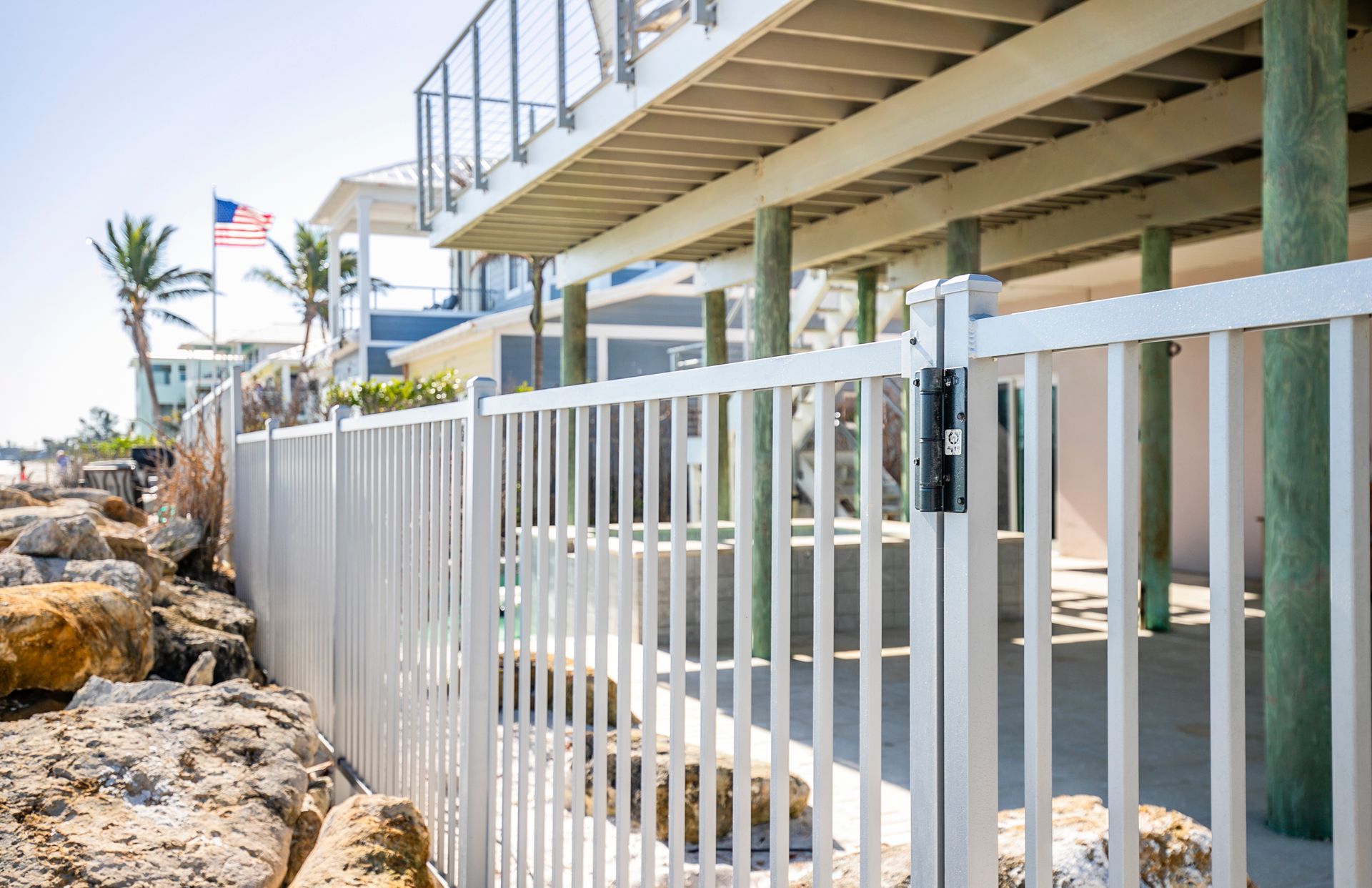 White metal fence by a house on pilings near a rocky beach with an American flag in the background.