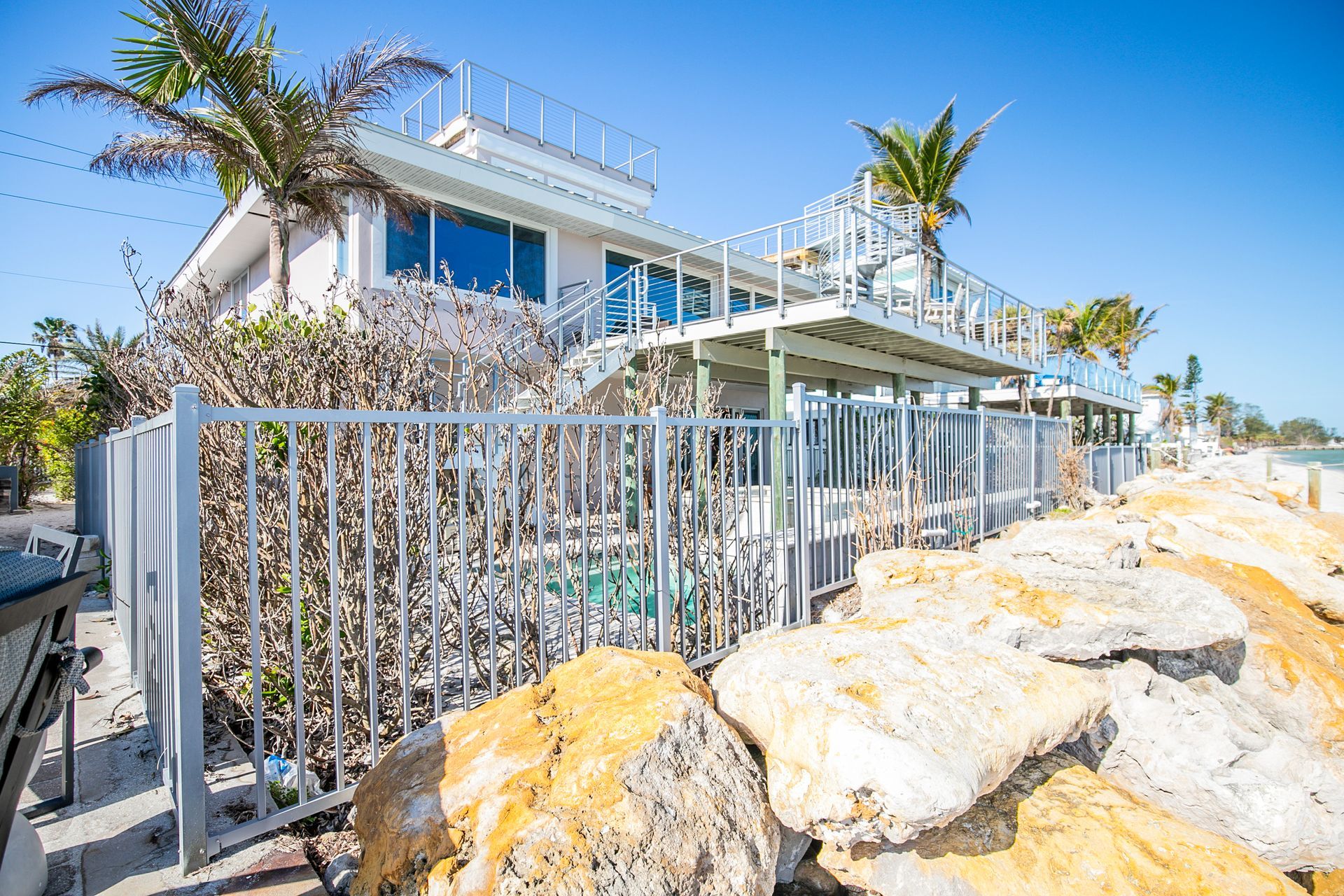 A beach house with a white exterior, raised wooden deck, and palm trees, partially hidden by a silver fence and large rocks.