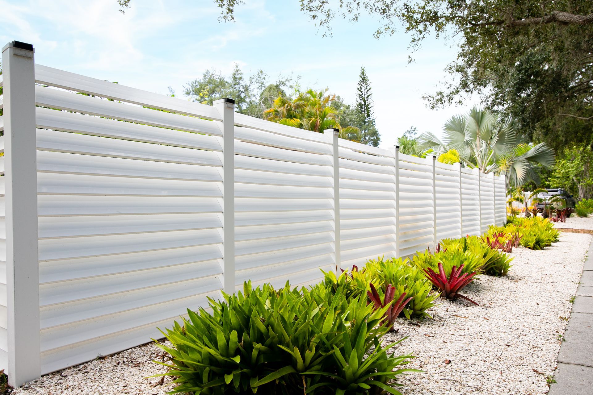 A long white horizontal-slat fence stands beside a landscaped bed with green plants and white gravel, outdoors.