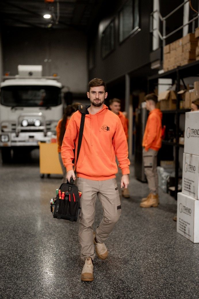A Man in an Orange Sweatshirt is Carrying a Tool Bag in a Warehouse — John McEwan Electrical In Dapto, NSW