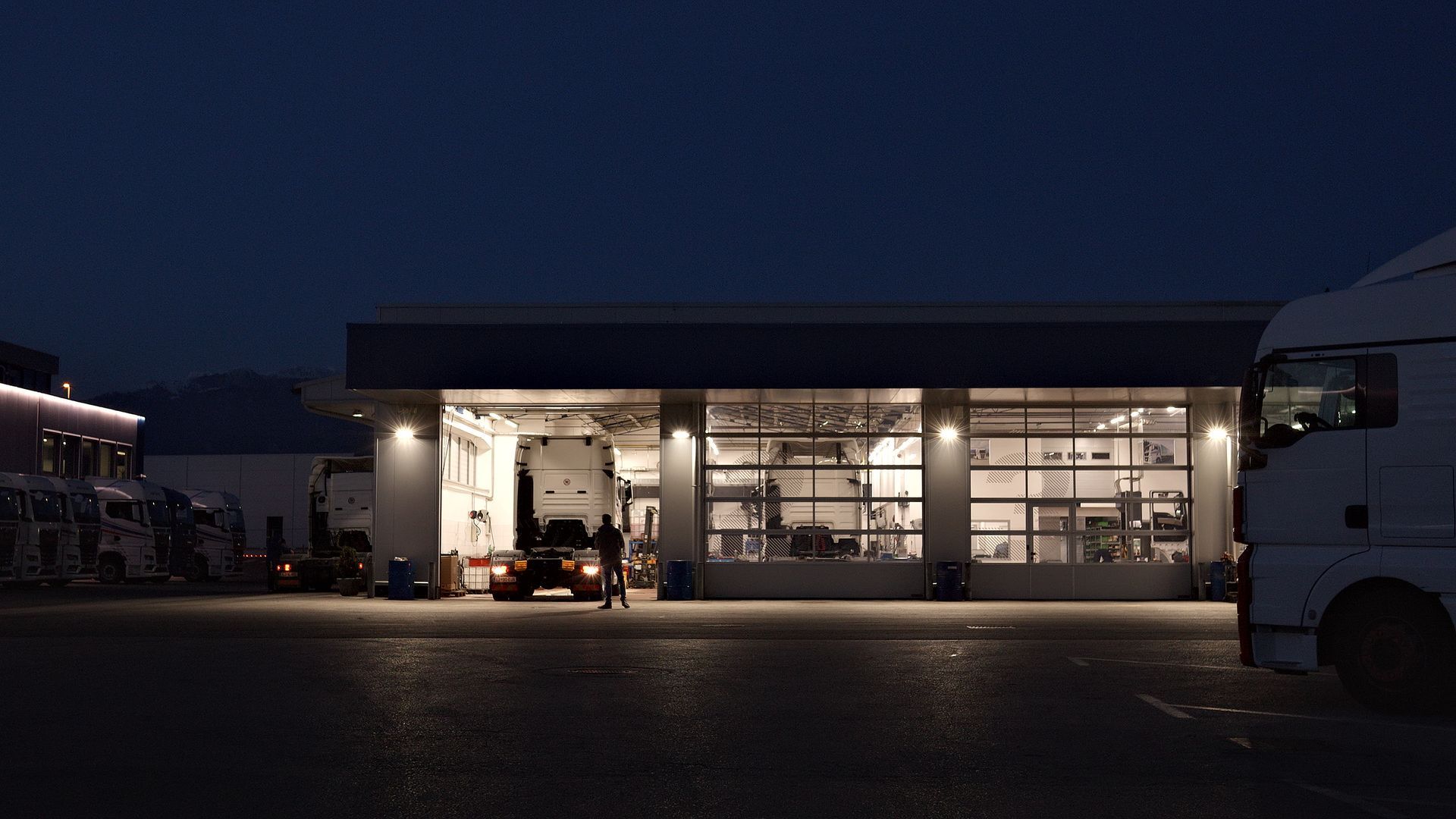A mechanic works late in a well-lit truck repair shop, surrounded by parked trucks. A mechanic works late in a well-lit truck repair shop, surrounded by parked trucks.