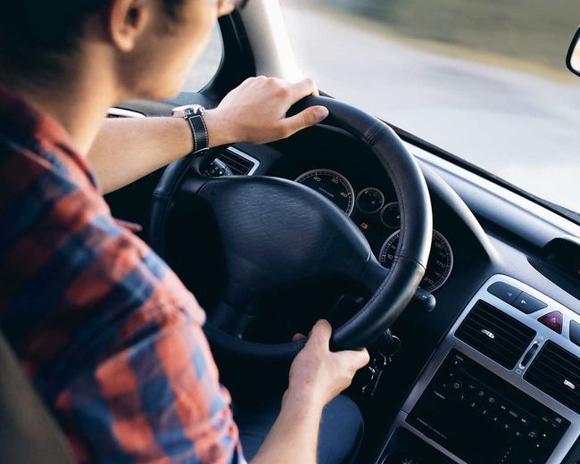 Driver holding steering wheel inside a car, wearing a red plaid shirt