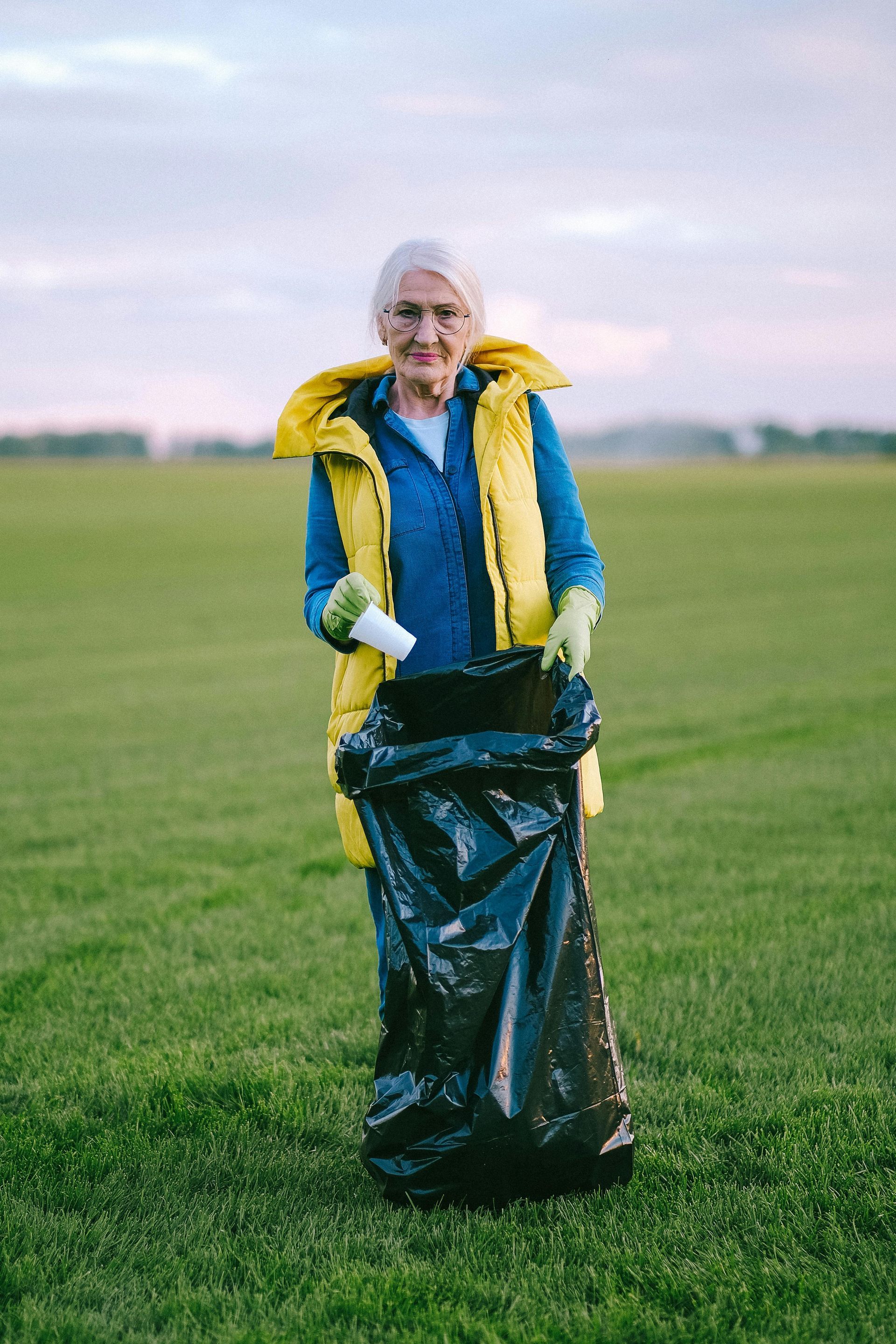 An elderly woman is standing in a field holding a trash bag.