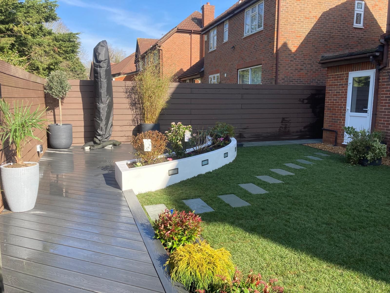A landscaped backyard with a stone path leading to a curved flower bed, bordered by a gray deck and green lawn. Houses are in the background.