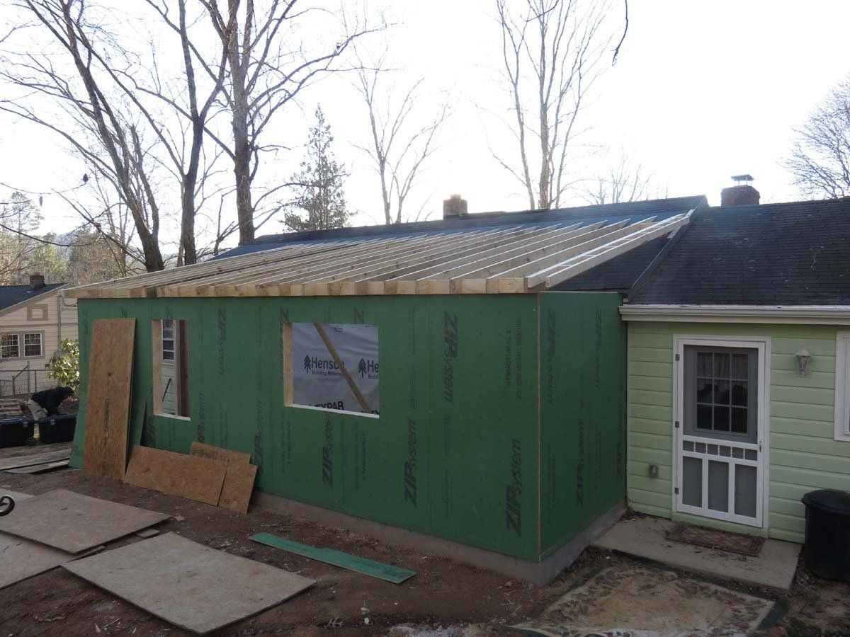 Green addition under construction beside a house, with roof framing and plywood panels in a yard.