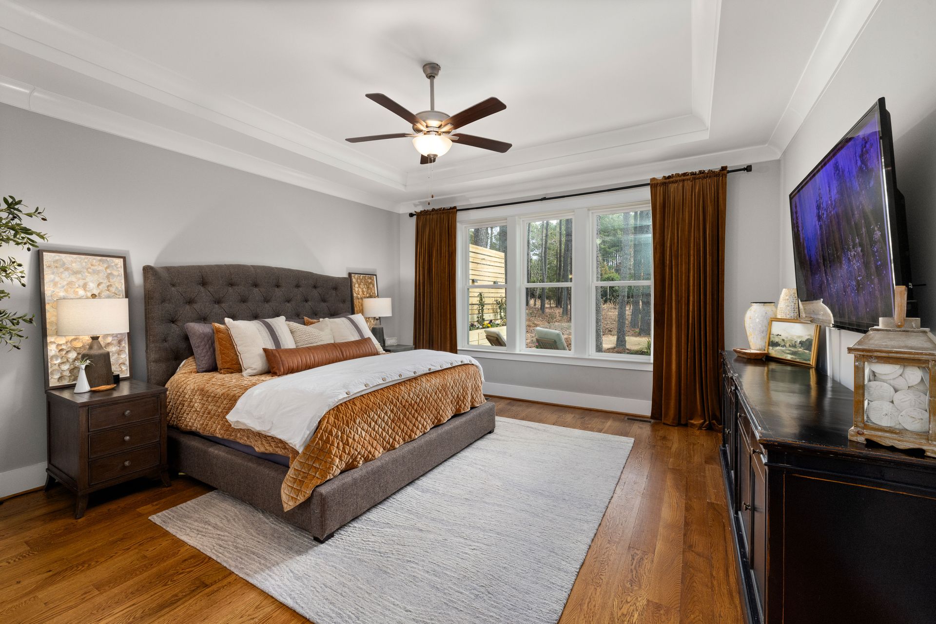 Bright bedroom with gray tufted bed, white rug, wood floors, large windows, and wall-mounted TV.