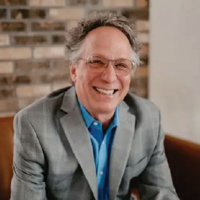 A man in a suit and glasses is smiling while sitting on a couch.