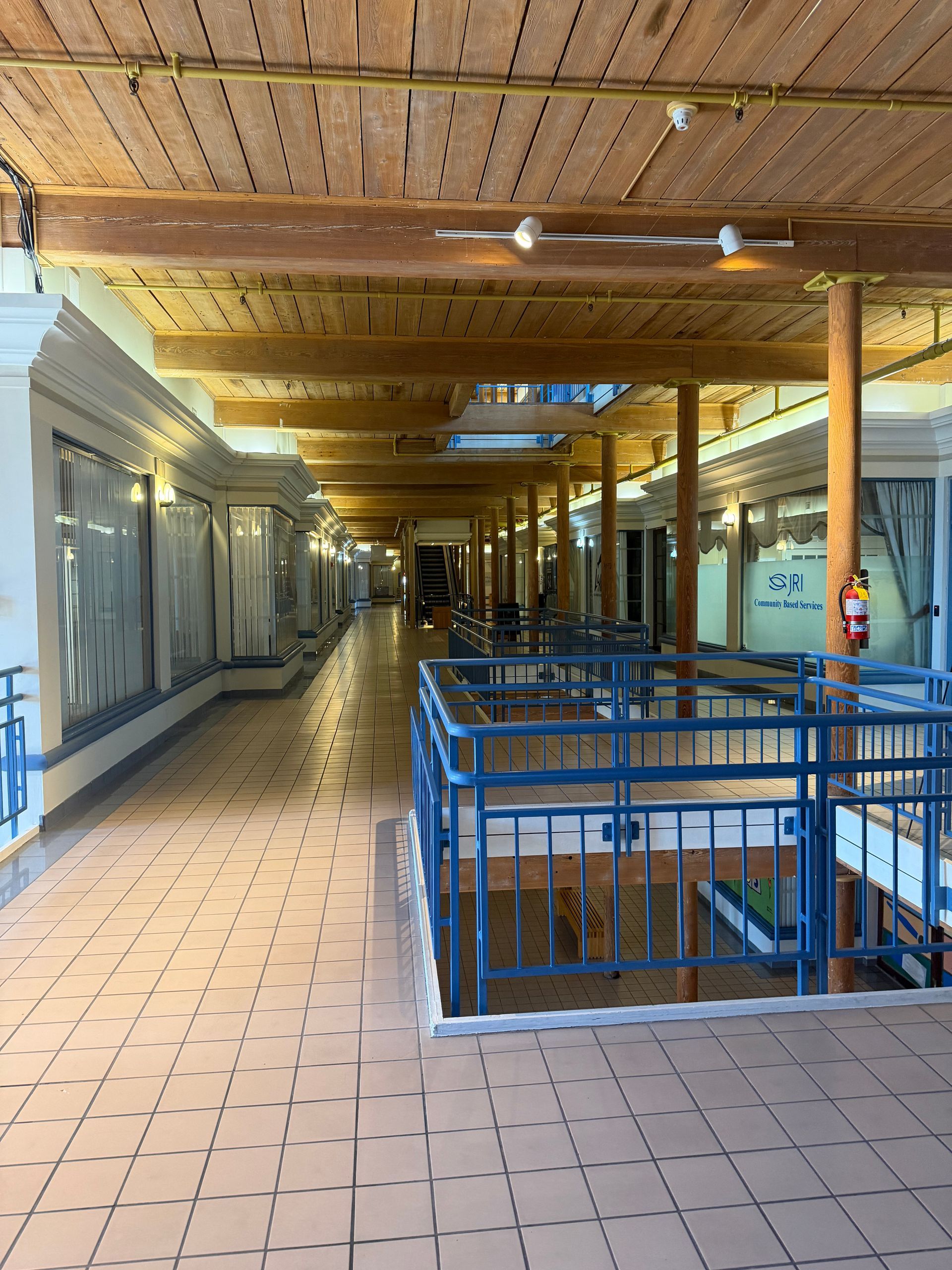 Covered corridor with tiled floor, blue railings, wooden ceiling, and closed storefronts leading into the distance