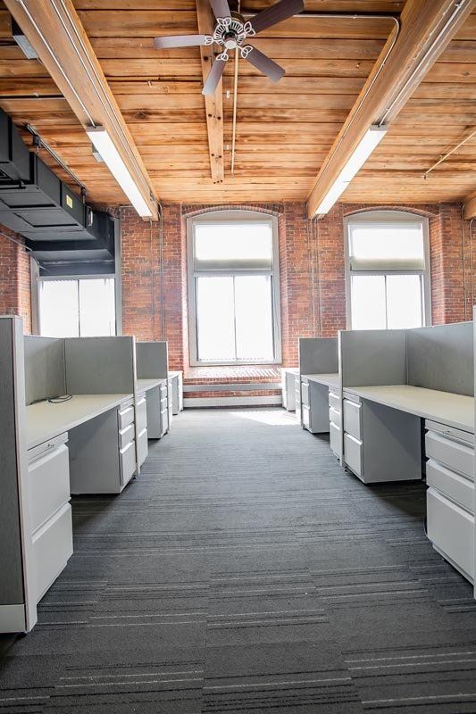 Open office with gray cubicles, brick walls, wood ceiling, and large windows.