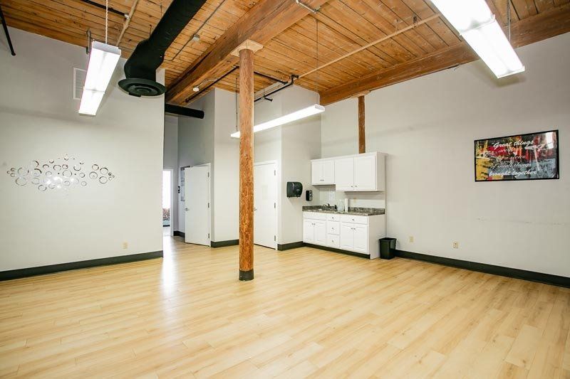 Empty loft-style room with light wood floors, exposed beams, kitchenette, and fluorescent lights