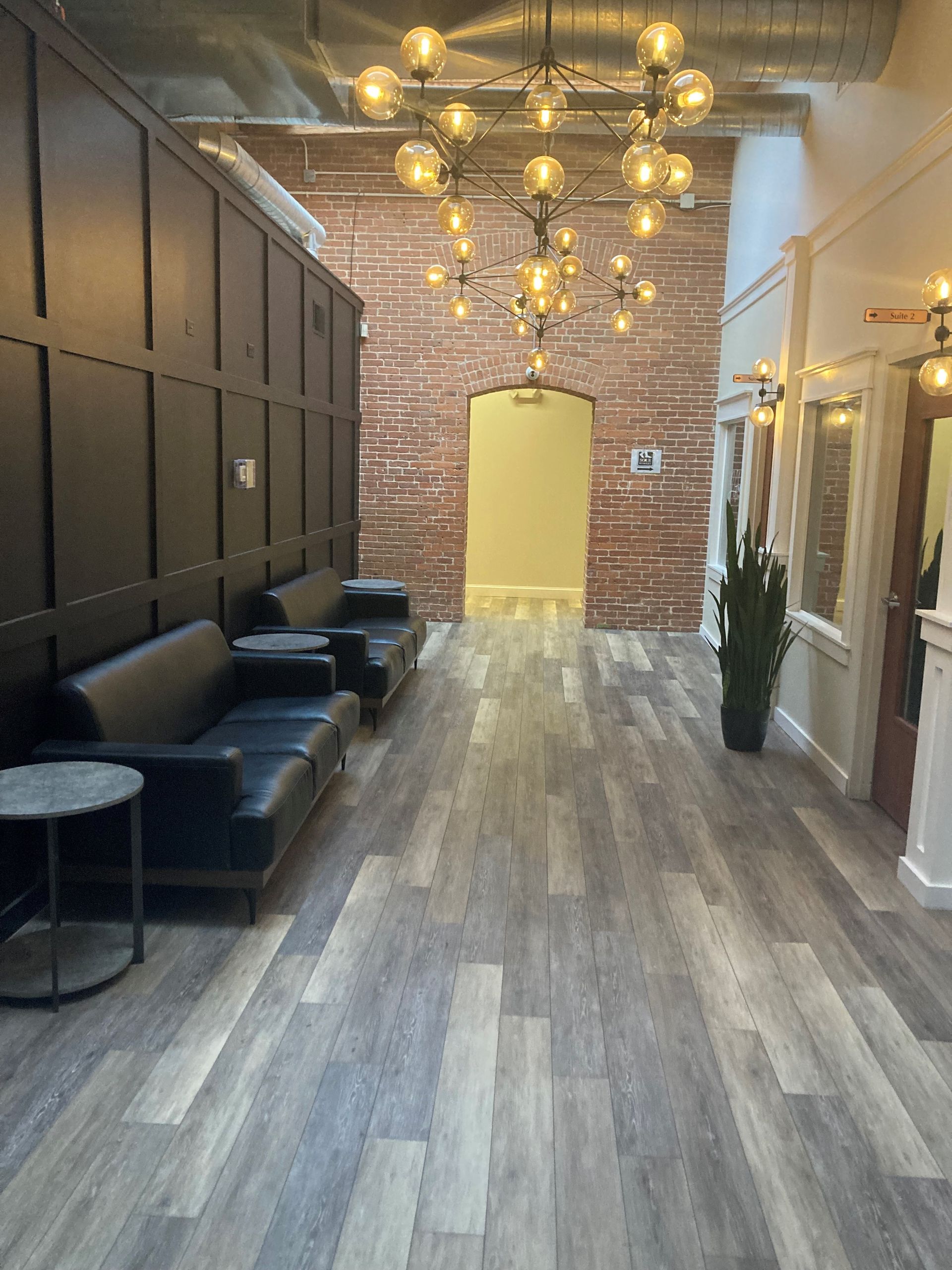 Stylish hallway with black seating, brass chandeliers, wood floors, exposed brick, and a yellow archway