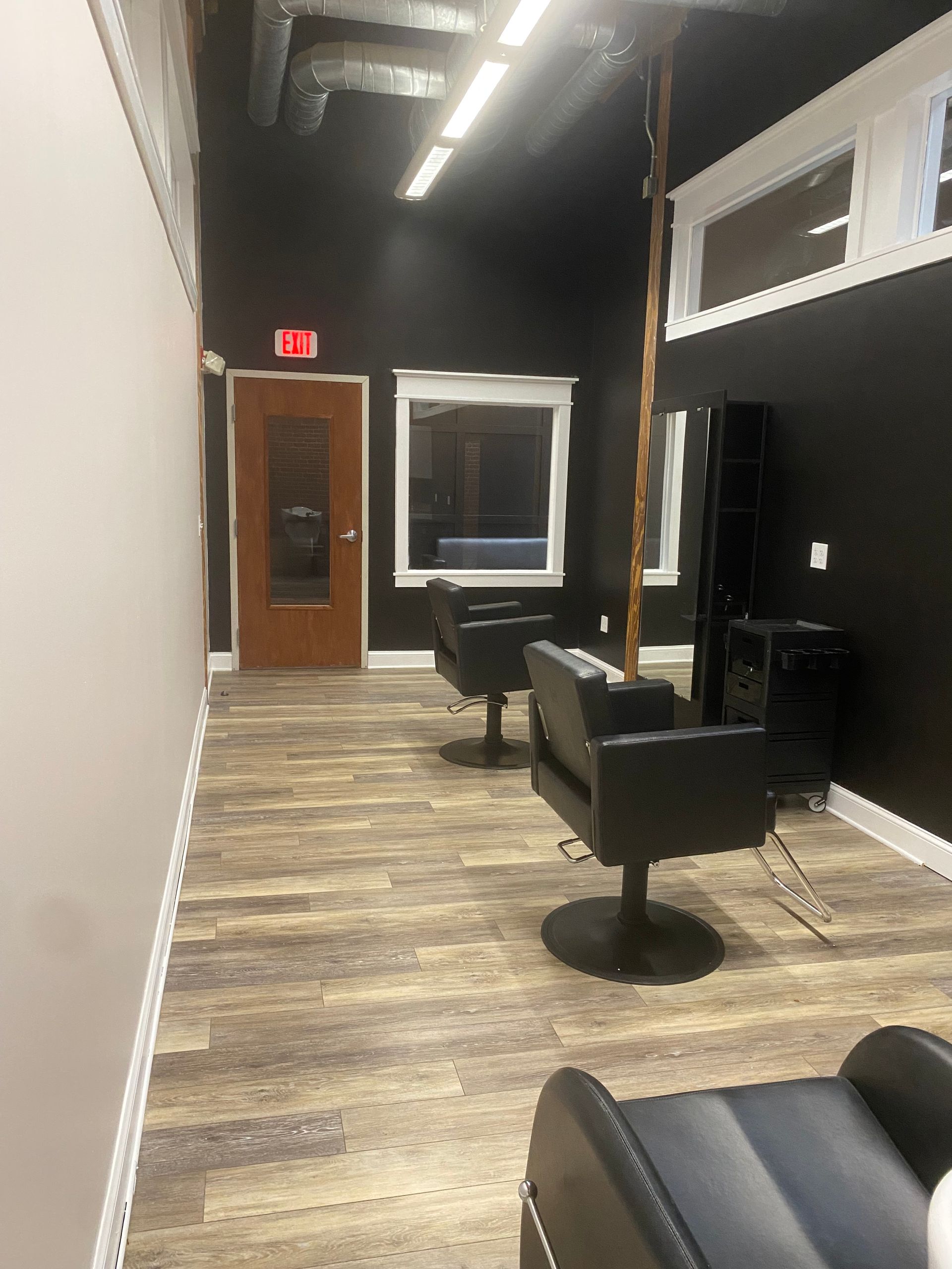 Narrow salon with black walls, wood-look floor, styling chairs, mirror, and an exit door at the far end