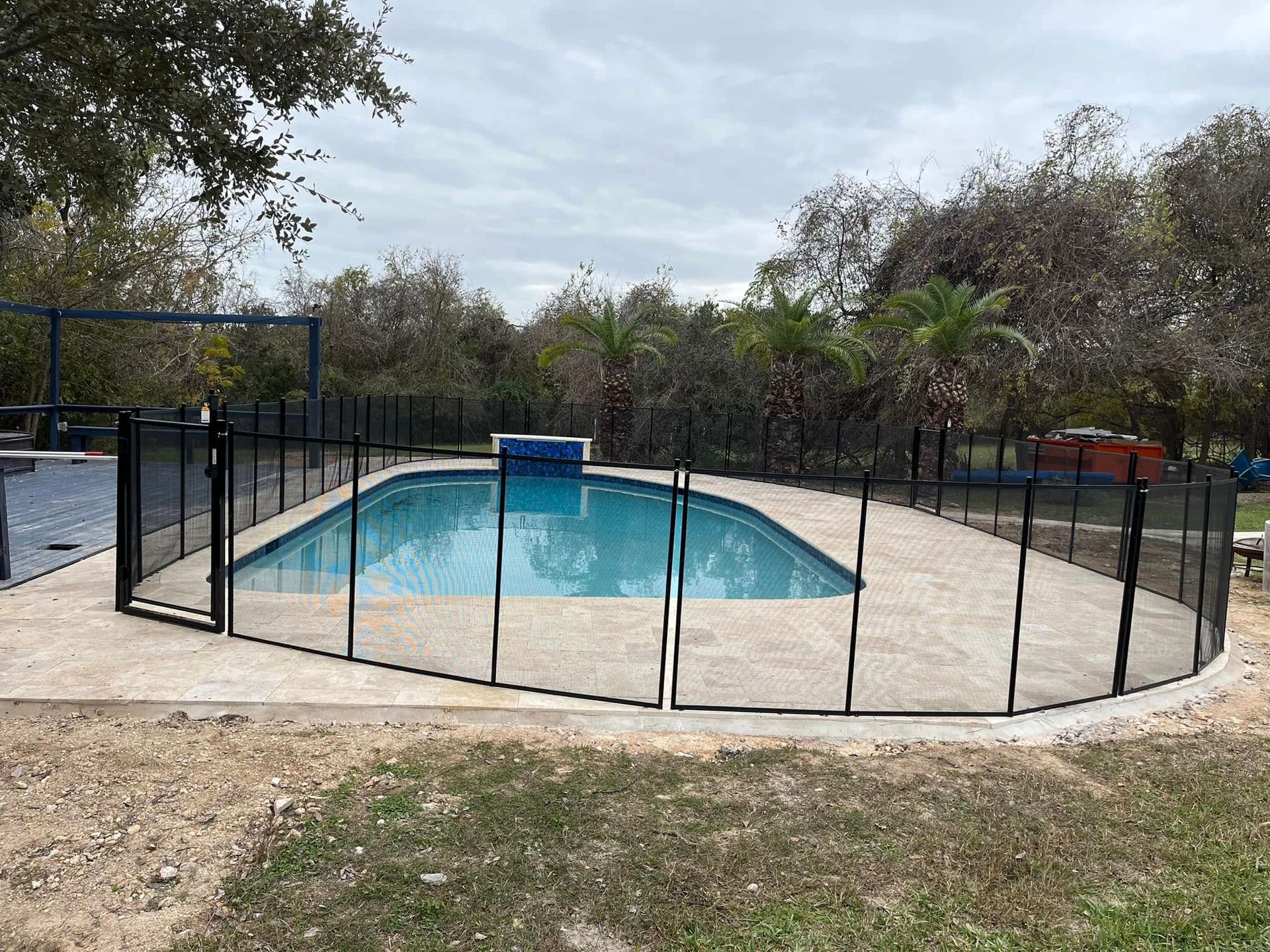 Backyard swimming pool enclosed by a black metal fence, with trees and cloudy sky in the background