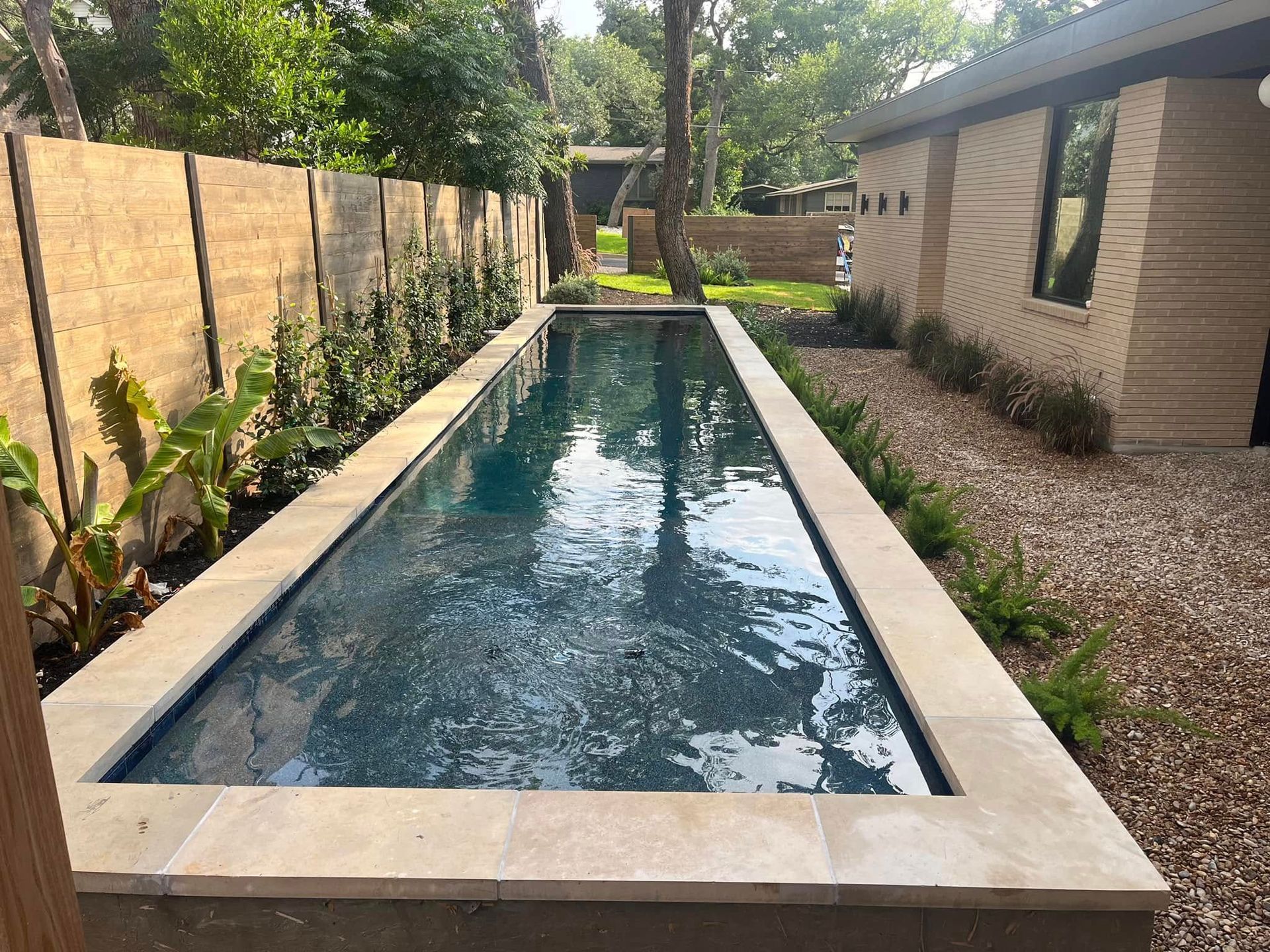 Long narrow backyard swimming pool beside a wood fence and modern house, surrounded by greenery.