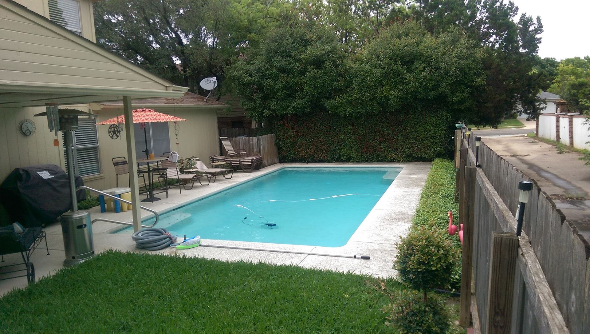 Backyard swimming pool beside a patio, grass, and wooden fence with lounge chairs nearby.
