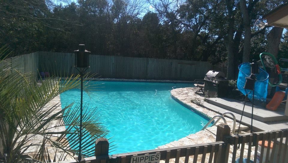 Backyard swimming pool with turquoise water, patio chairs, and trees behind a wooden fence
