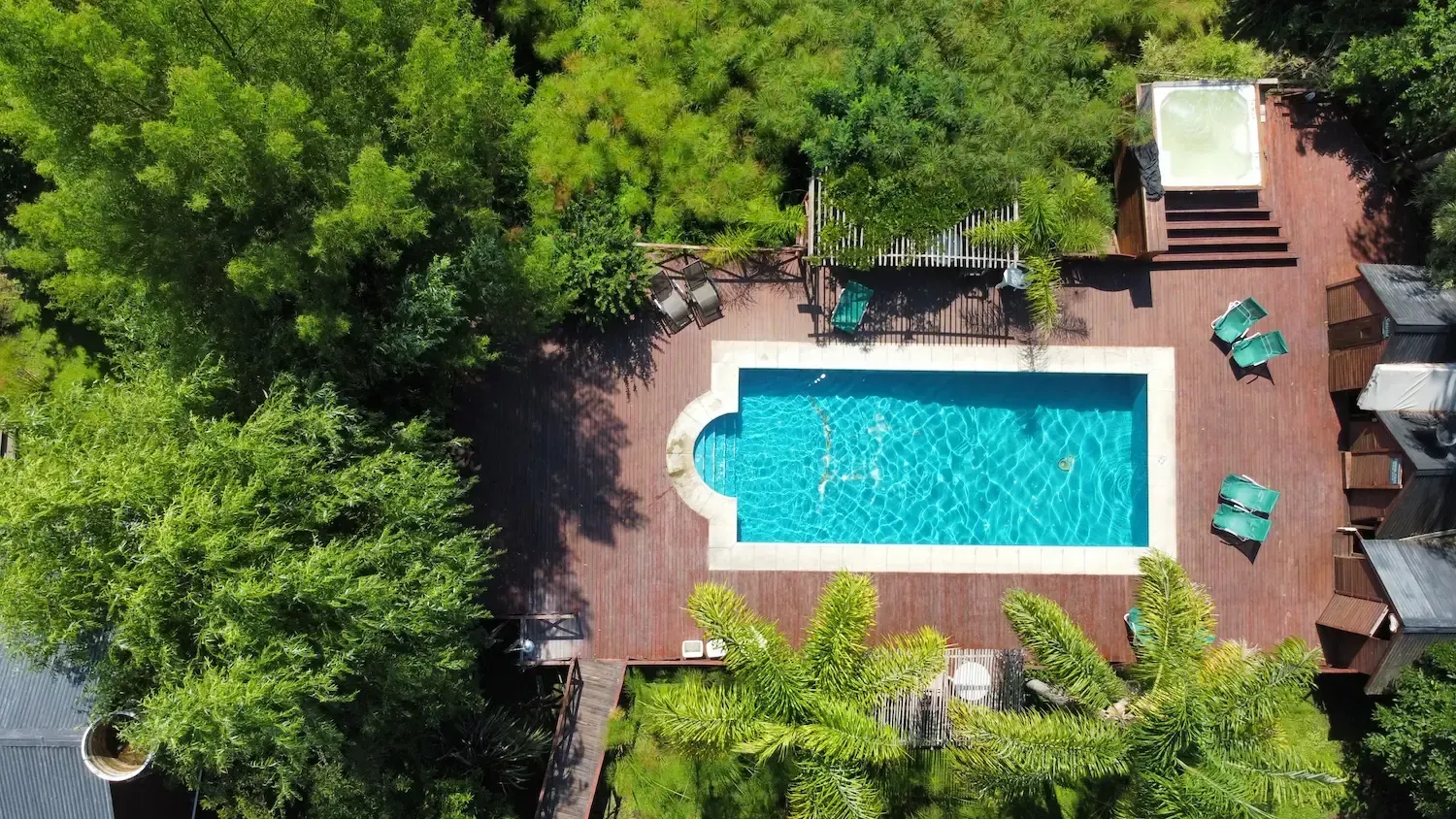 Vista aérea de una piscina rectangular rodeada por una terraza de madera, árboles y pequeños edificios.