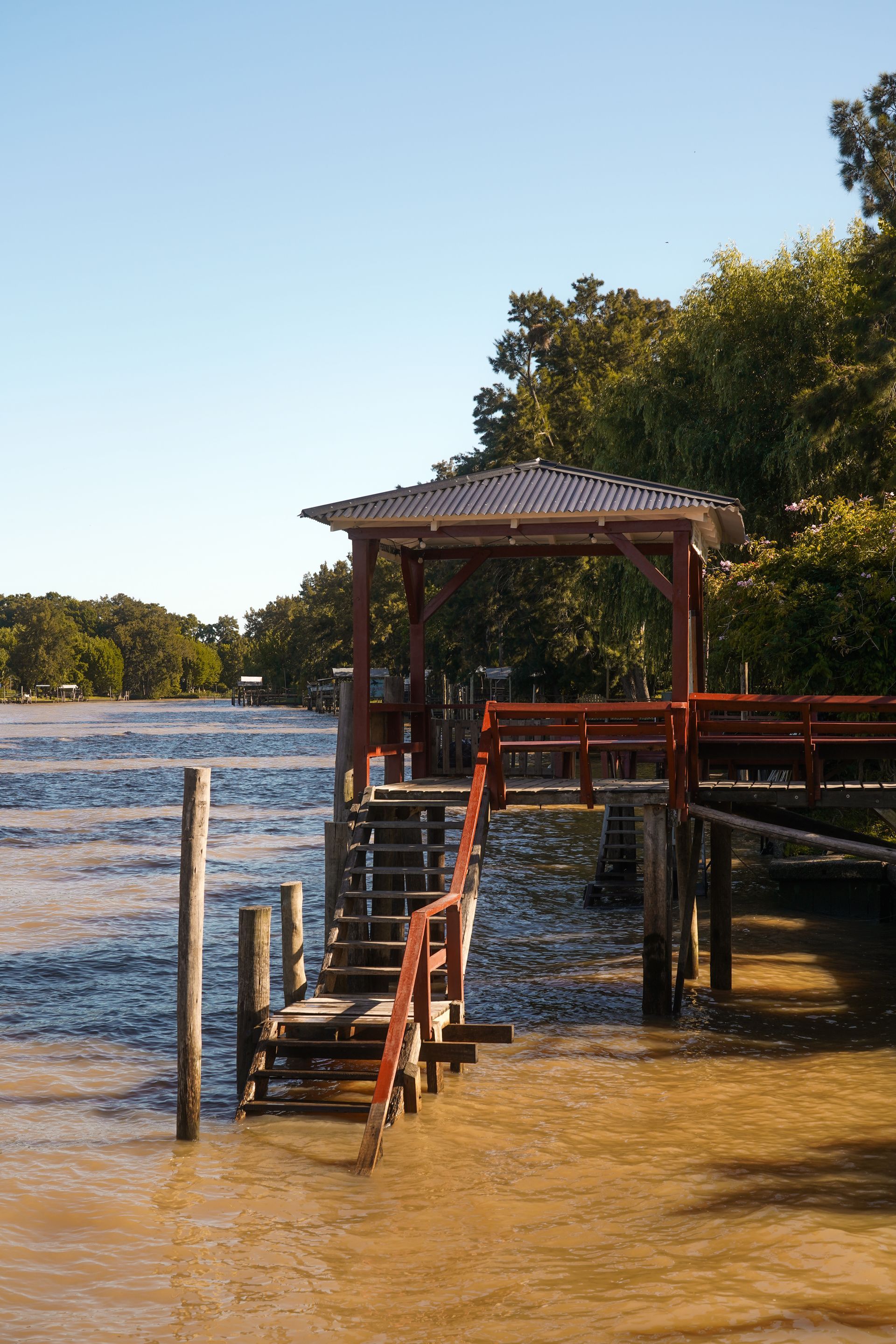 Muelle de madera con escaleras y mirador sobre aguas turbias. Árboles bordean la orilla y un cielo despejado.