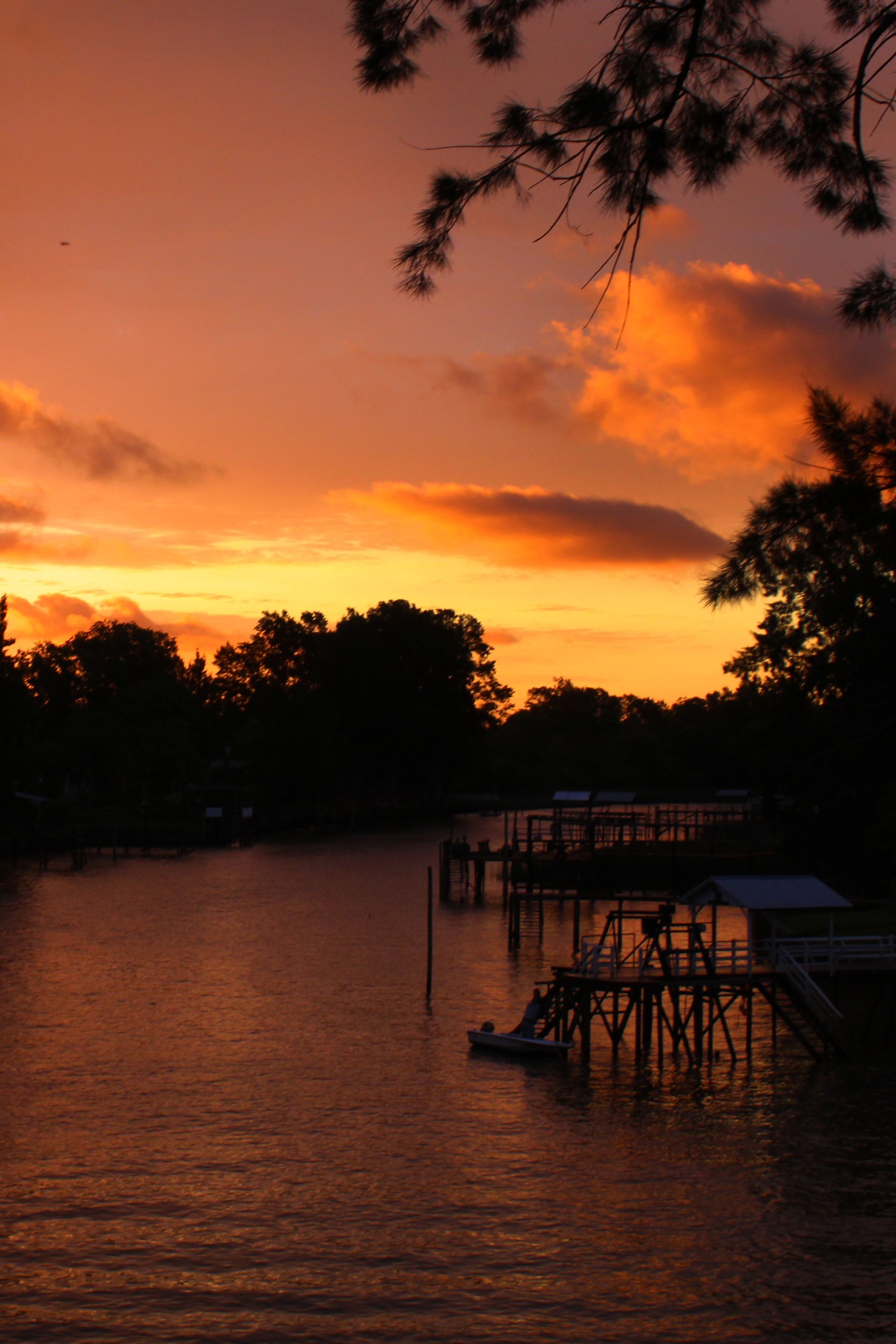 Atardecer sobre un río tranquilo con muelles; cielo naranja y amarillo, siluetas oscuras de árboles y embarcaderos.