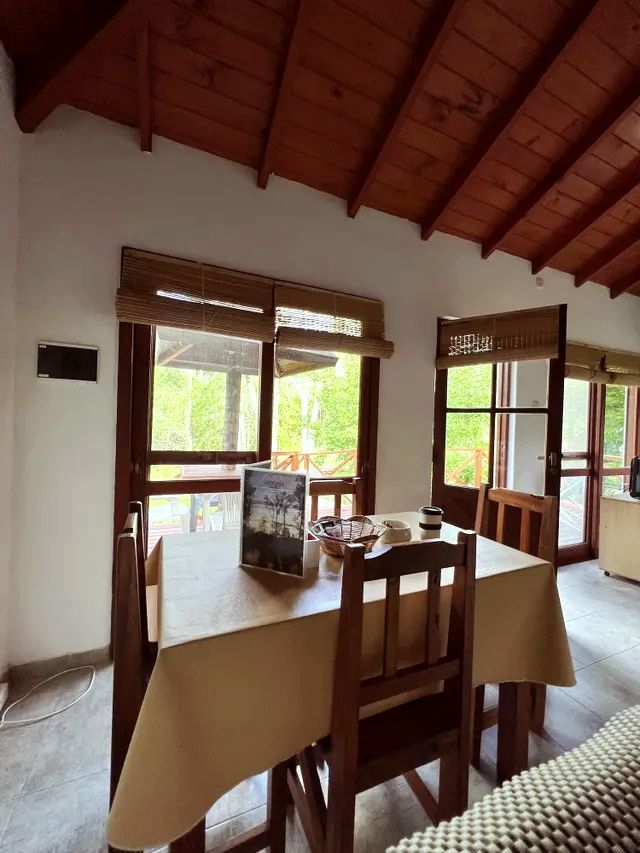 Comedor con mesa y sillas de madera, ventanas y persianas de mimbre. Mantel color crema. Vigas en el techo.