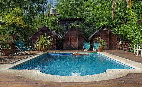 Piscina rodeada por una terraza de madera, pequeñas cabañas y exuberante vegetación. Dos sillas se encuentran junto a la piscina.