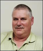 A man with a mustache is wearing a green shirt and smiling.