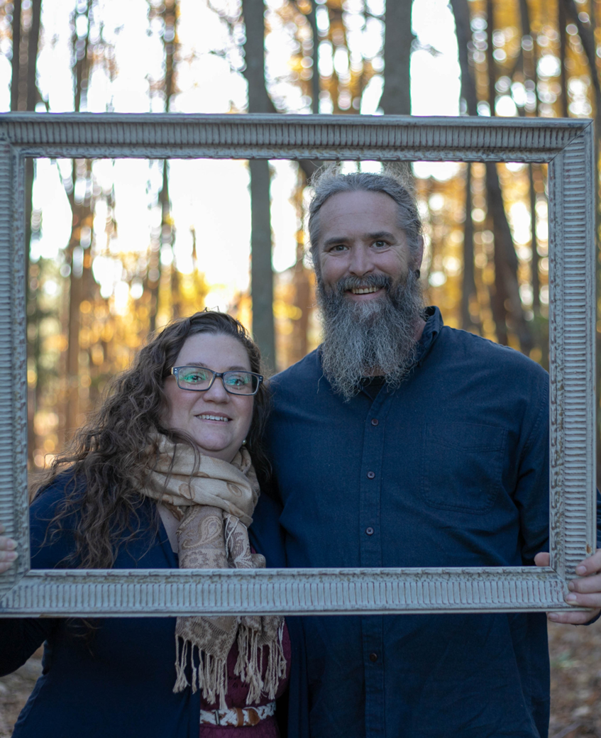 A man and a woman are holding a picture frame in the woods.