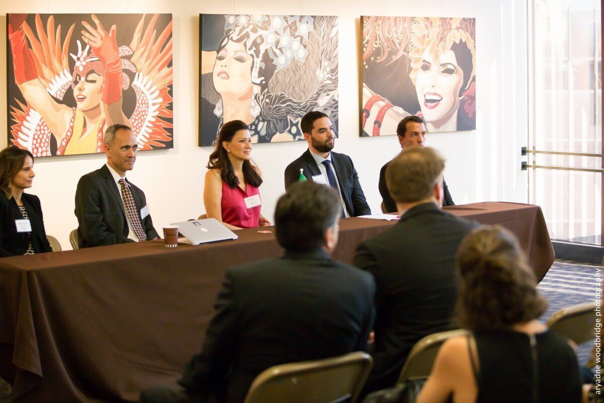 Panel of speakers at a conference, seated at a table, in front of artwork; audience in foreground.
