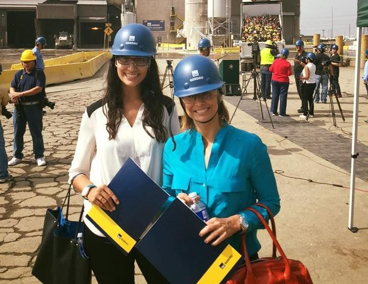 Two women smiling, wearing hard hats, holding folders; industrial setting, people in the background.