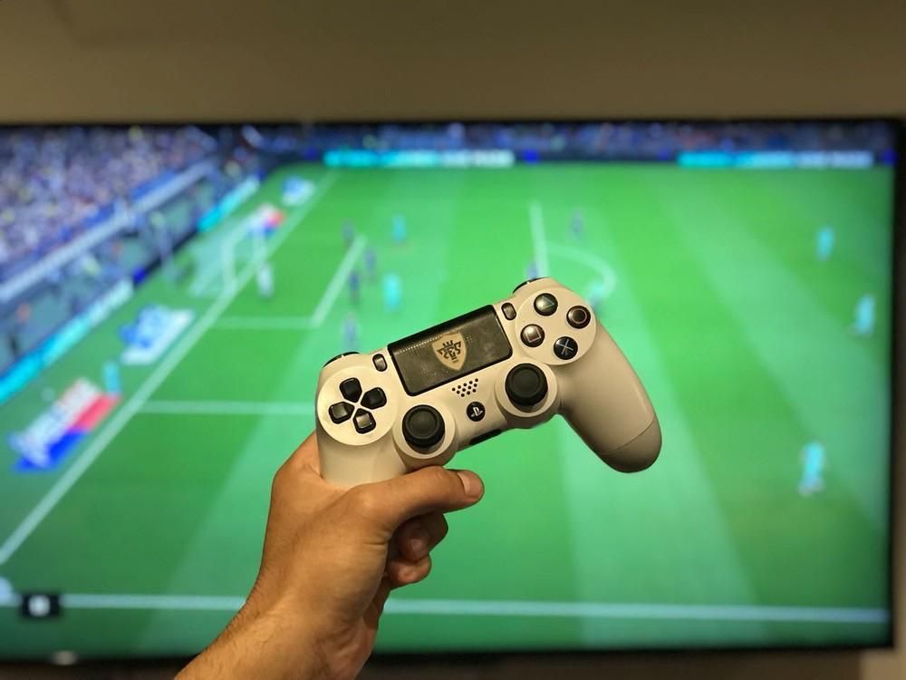 Hand holding a white PlayStation 4 controller with a soccer game on a TV screen in the background.