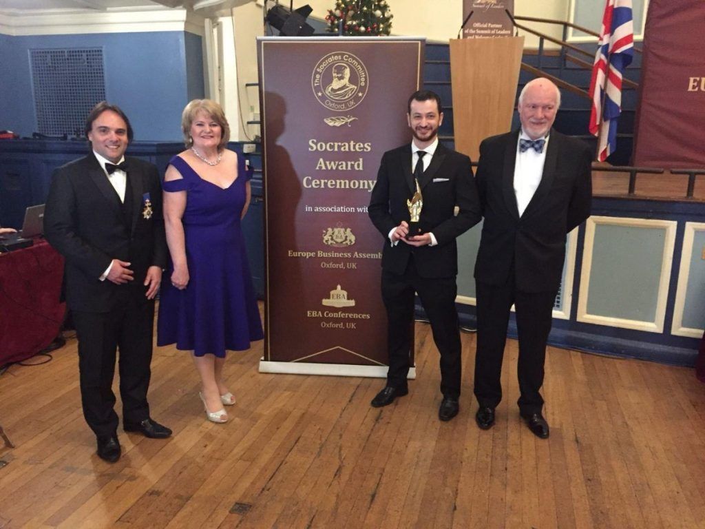 Four formally dressed people pose near a banner, one holding an award. Interior setting.