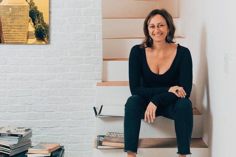 Woman with brown hair sits on stairs, smiling. Wearing a black top and jeans. White brick wall and artwork behind her.