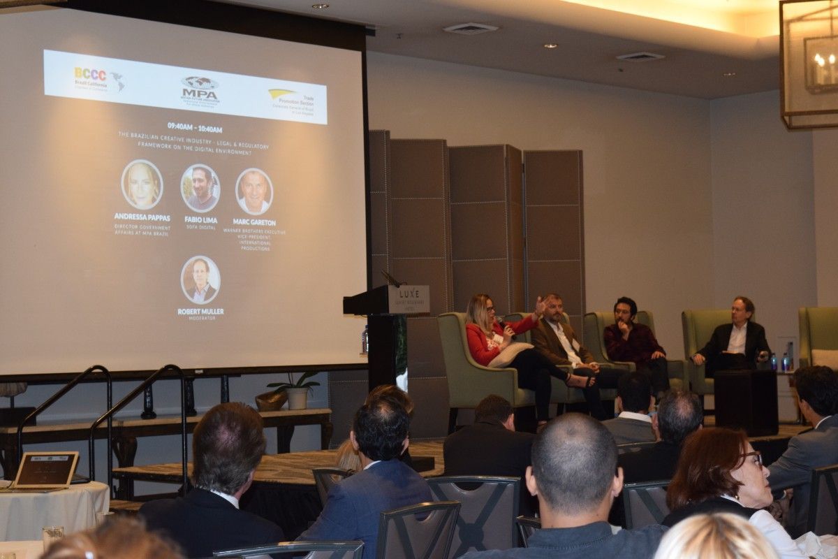 Panel discussion at a conference with speakers on stage, audience seated, projector screen with speakers' names.