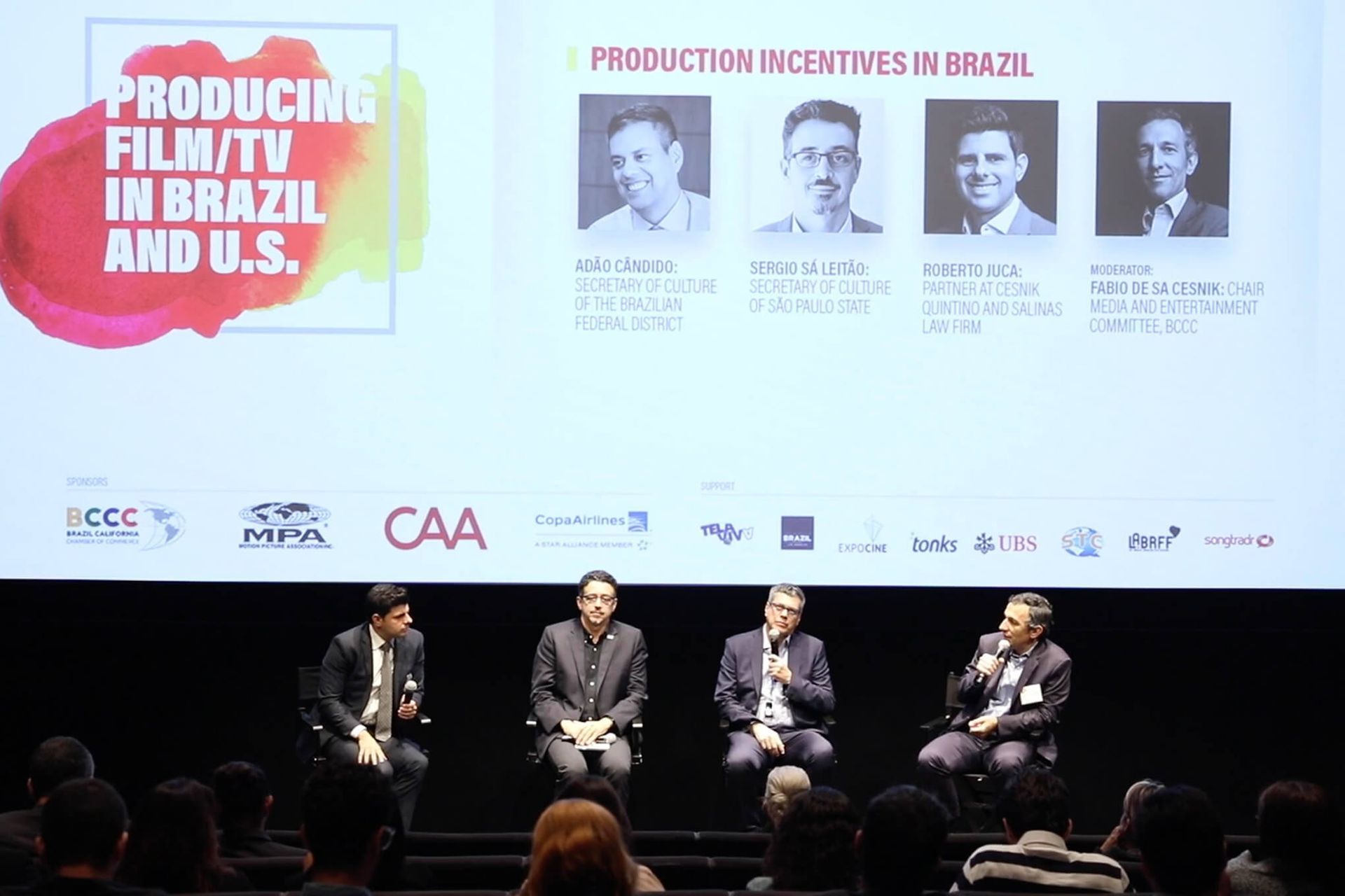 Panel discussion: four men on stage speaking, audience in front. Title: 