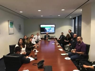 People seated around a conference table in a meeting. A screen shows a presentation.